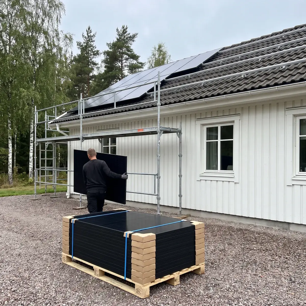 Pall med helsvarta Trina Vertex S+-paneler på uppfarten till en vit villa medan en montör bär en panel mot ställningen.