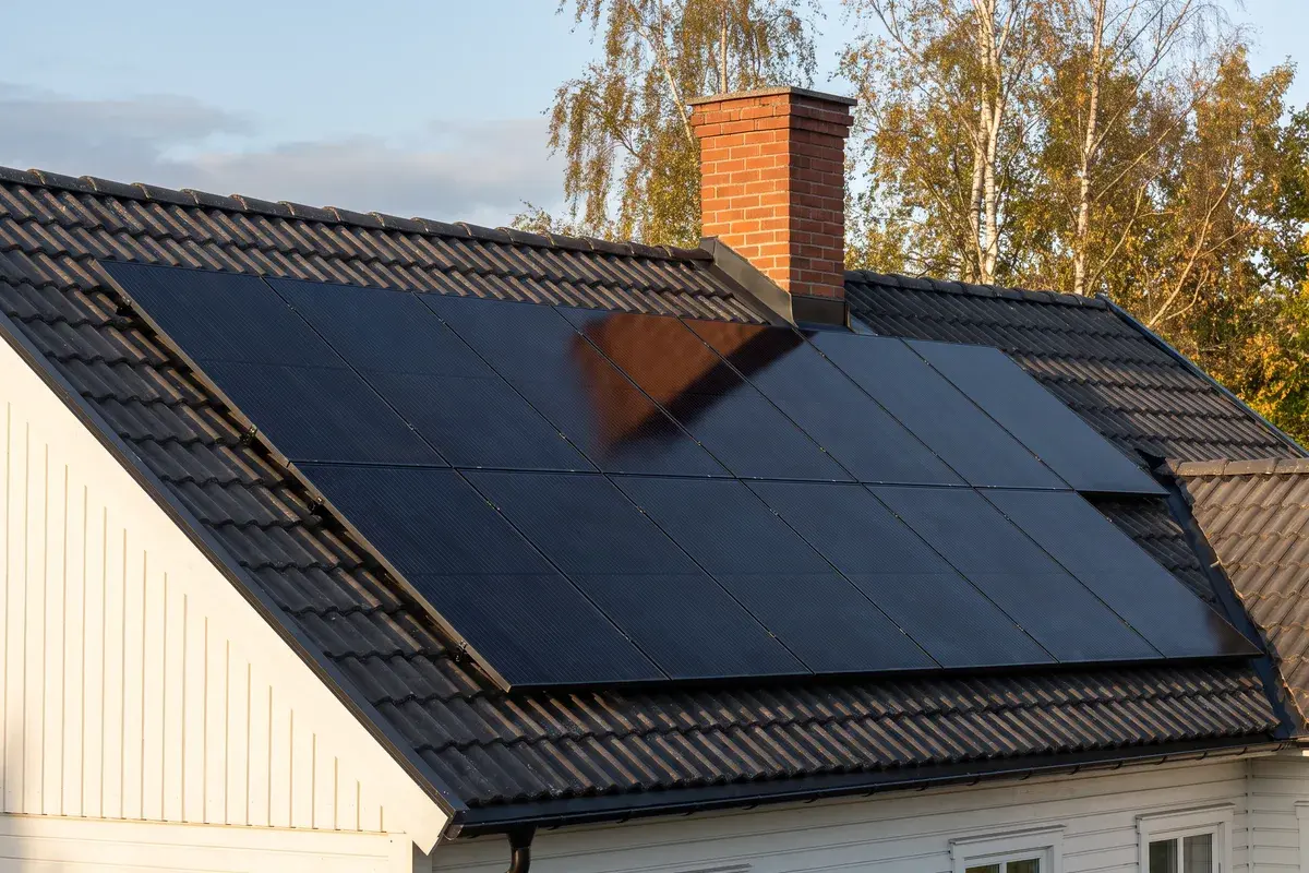 Skorstenens skugga faller snett över tre helsvarta Trina Vertex S+-paneler på ett betongpannetak en eftermiddag.