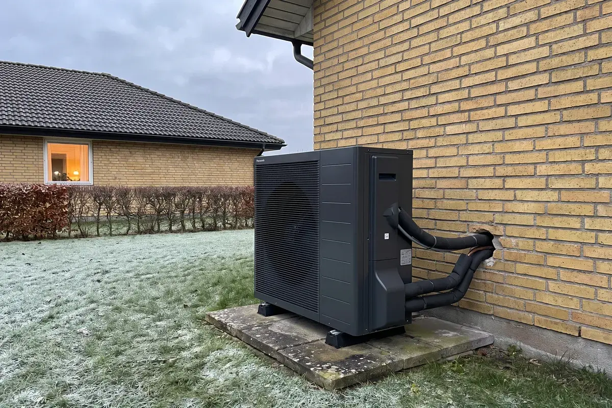 Panasonic Aquarea L luft-vattenvärmepump på markstativ vid en gul tegelvilla i Skåne en grå januaridag.