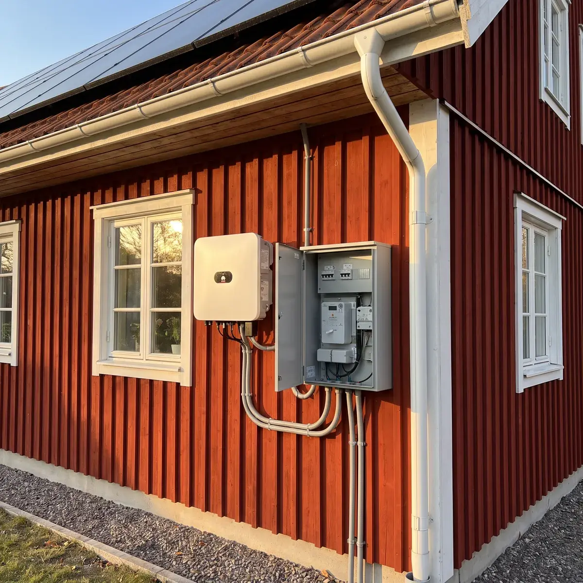 Huawei SUN2000-växelriktare och öppet mätarskåp på gaveln till ett falurött trähus med solpaneler på taket.
