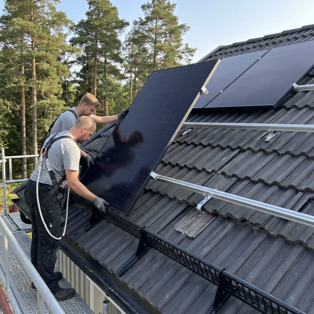 Två montörer på en ställning lägger en helsvart REC Alpha Pure-RX-panel på K2-skenor över ett mörkt betongpannetak.